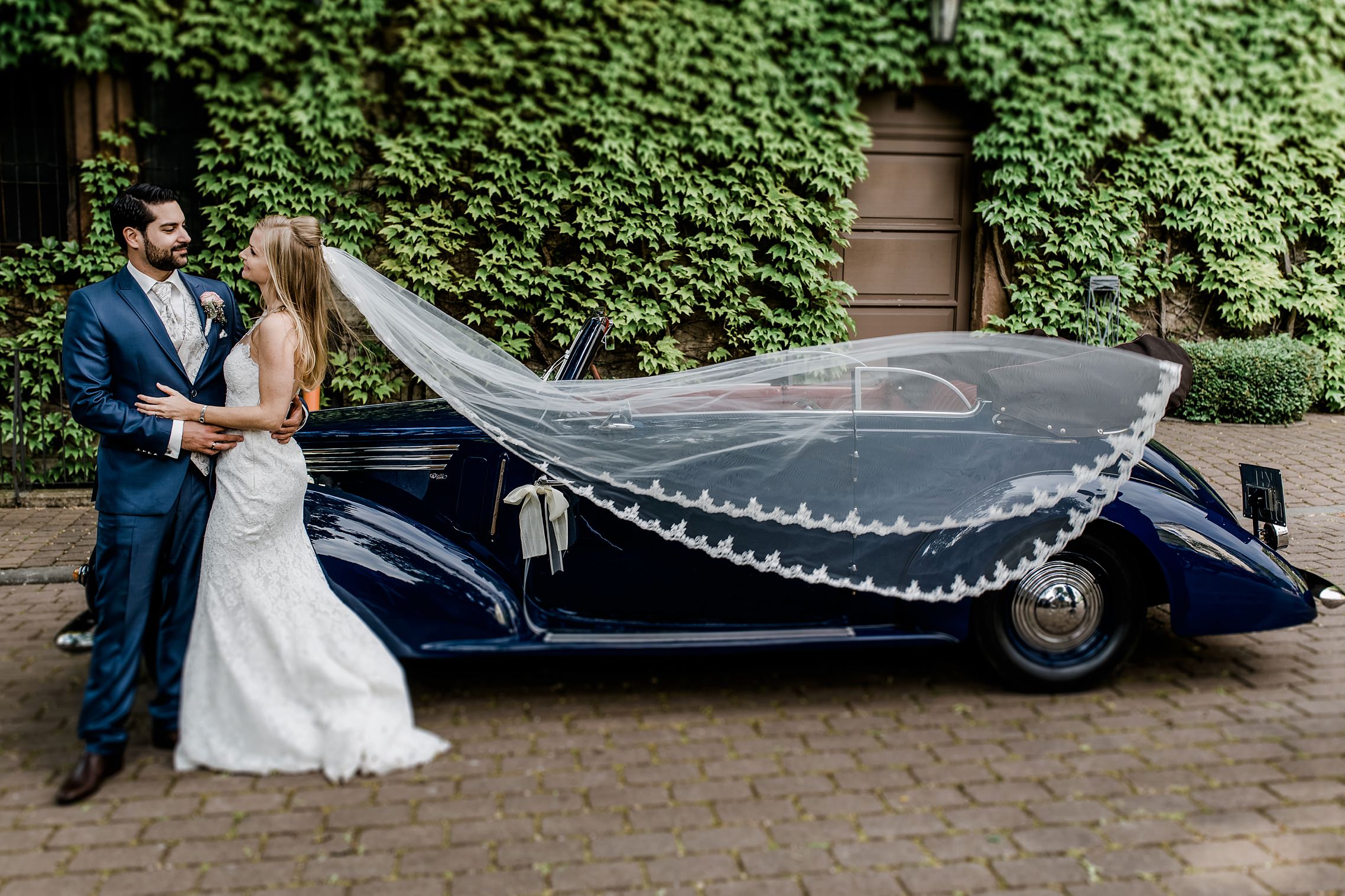Hochzeitspaar mit fliegendem Schleier vor einem Fiat-Oldtimer von 1930