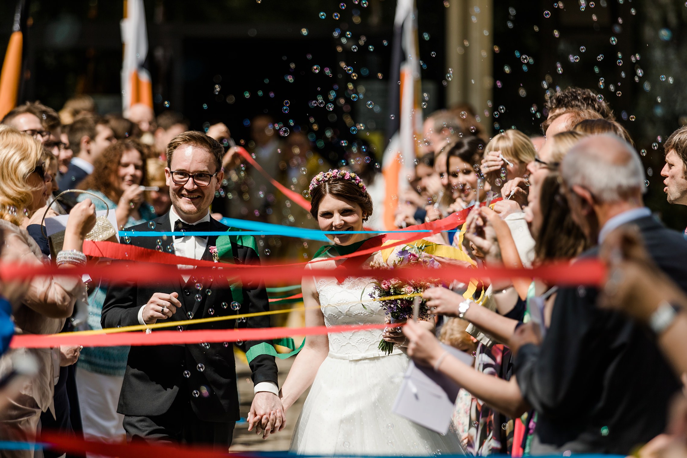 Brautpaar schreitet nach dem Gottesdienst durch ein Spalier aus Seifenblasen und Farbbändern.