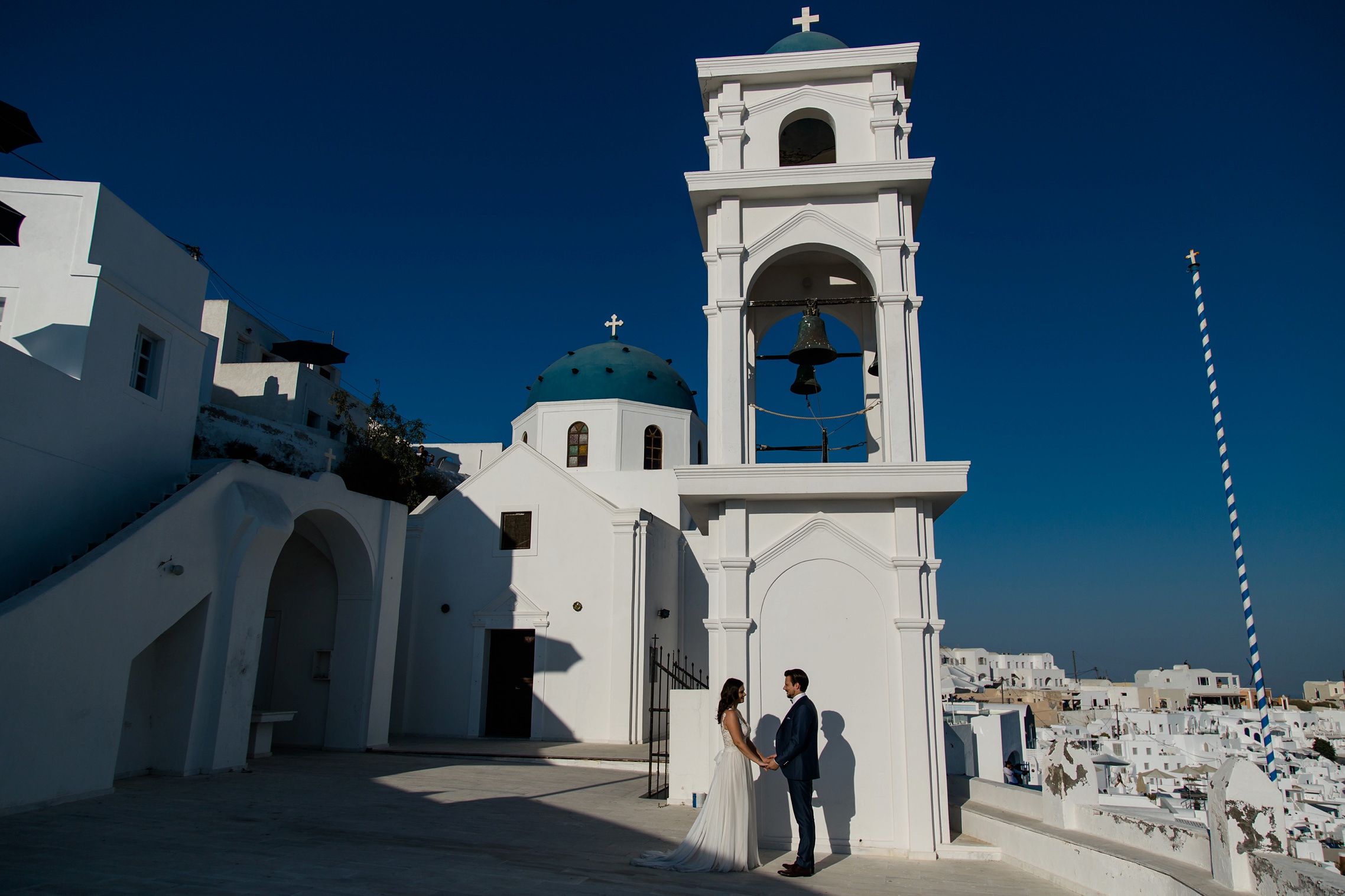 After-Wedding Santorini: Braut und Bräutigam stehen vor einer typischen Santorini-Kulisse in blau und weiß. Kirchturm und Kirchkuppel im Hintergrund.