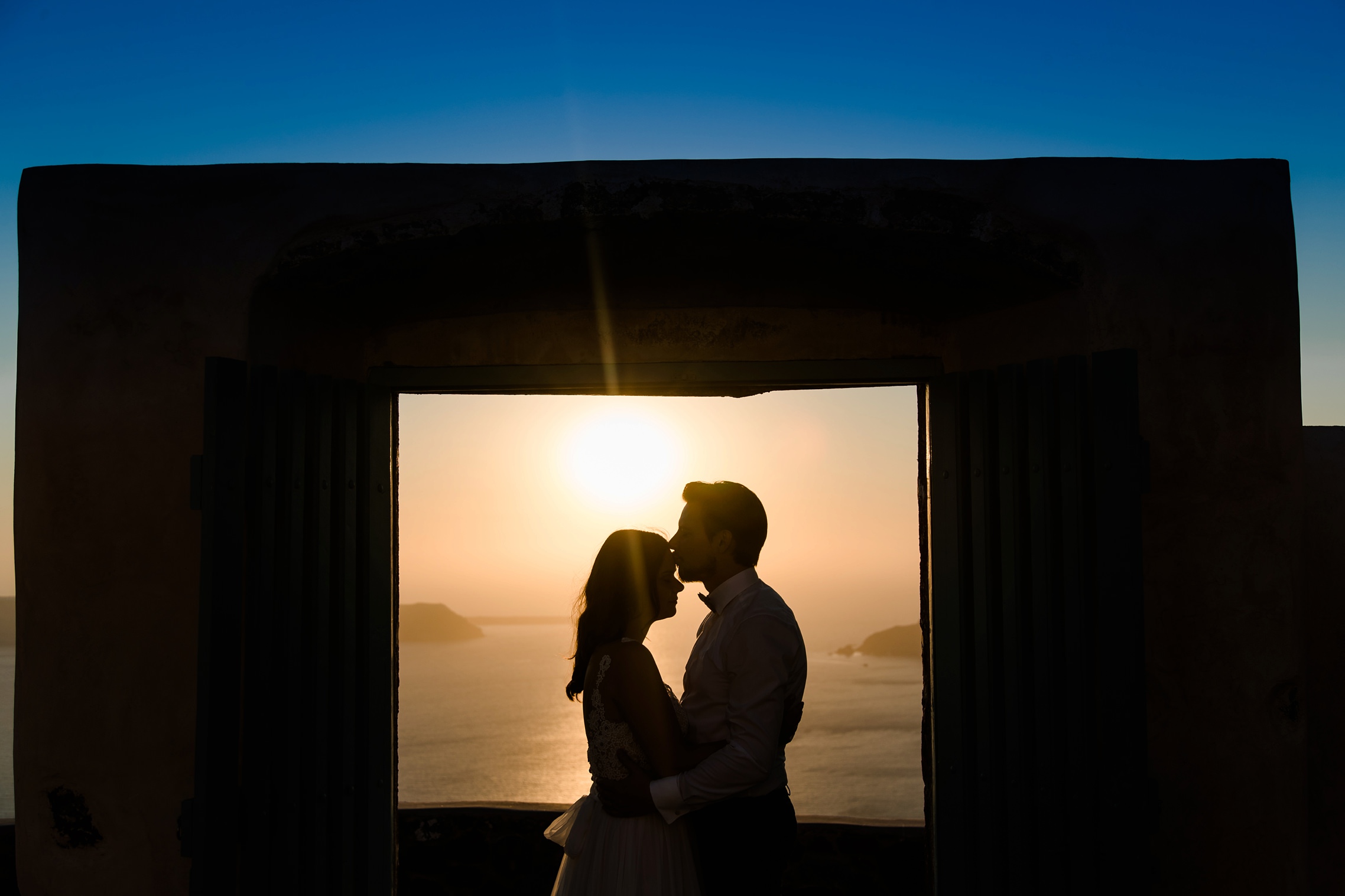 After-Wedding Santorini: Küssendes Hochzeitspaar beim Sonnenuntergang vor einem blau-orangenen Himmel.