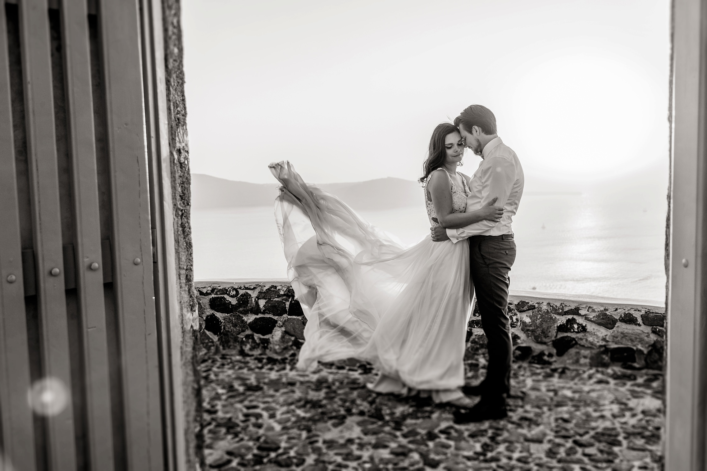 Foto in schwarzweiß: das Brautkleid fliegt in der Meeresbrise beim After-Wedding-Shooting auf Santorini.