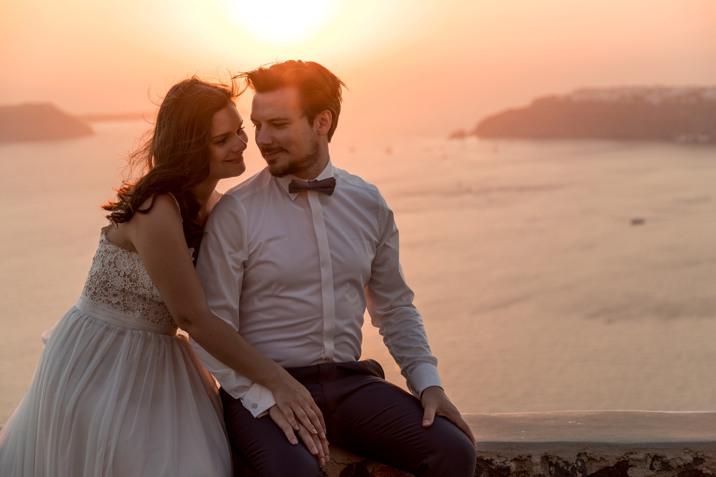 Romanisches Hochzeitspaar genießt den Sonnenuntergang auf Santorini beim After-Wedding Shooting und sitzt auf einer Steinmauer.