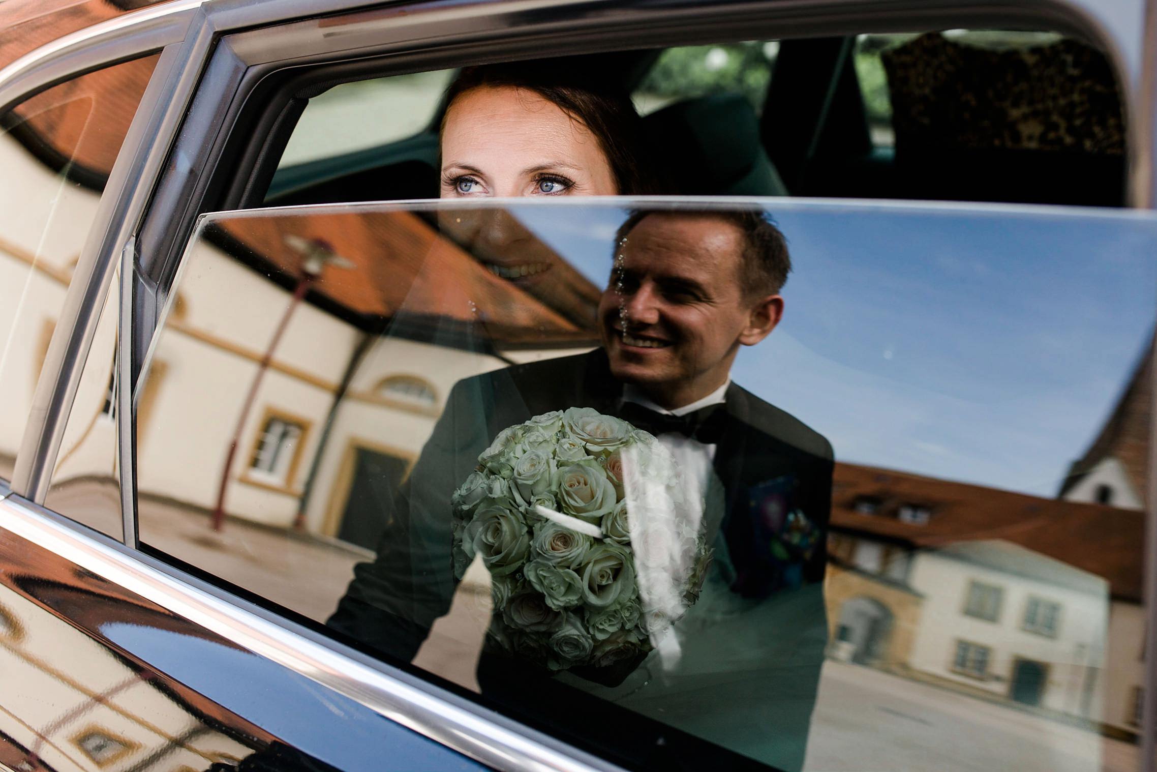 Bräutigam spiegelt sich in Autoscheibe, Braut schaut ihn verliebt an.