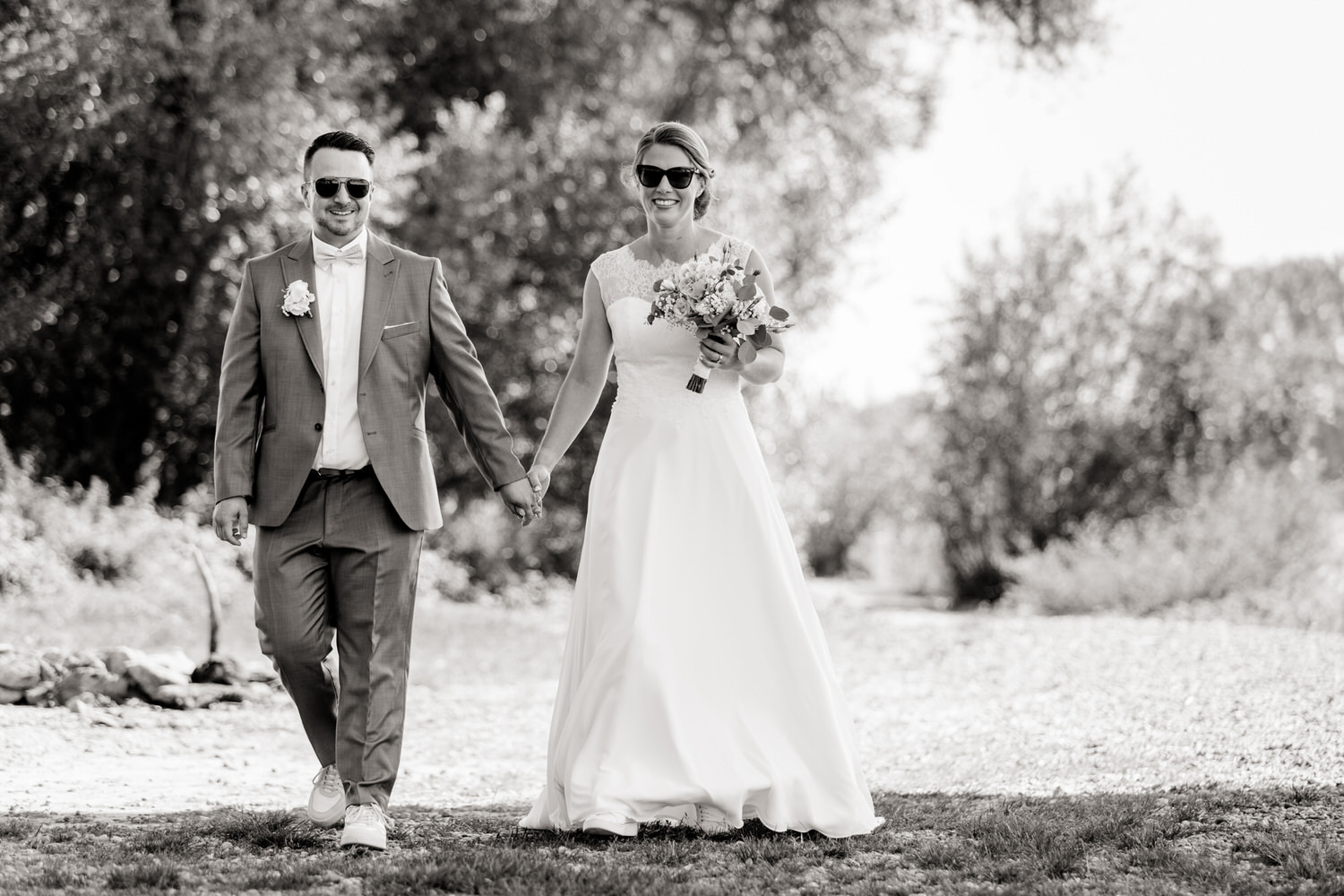 Cooles schwarzweiss Bild: Hochzeitspaar schlendert mit Sonnenbrillen in der Natur.